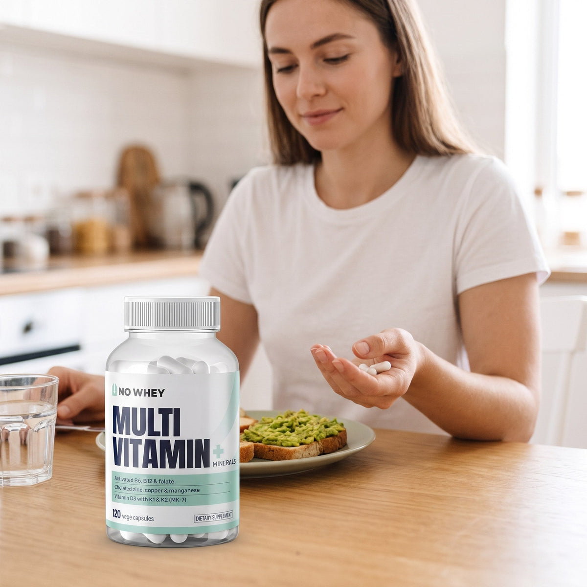 Woman at a kitchen table at breakfast holding two capsules, a bottle of NoWhey Multivitamin + Minerals beside her glass of water